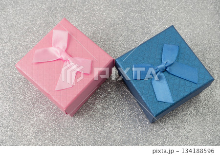 Two small boxes with ribbons are placed on a shiny surface. One box is blue and the other is pink. These boxes are ready for gifting during a party or event. 134188596