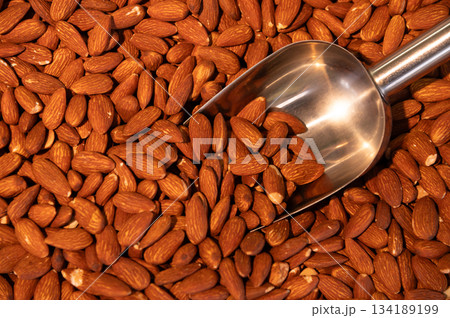 A large quantity of roasted almonds are on display at the store. A stainless steel scoop is immersed in the almond kernels. 134189199