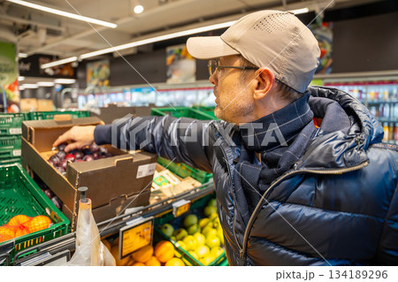 Man picking fresh plums in the fruit and vegetable section of a supermarket. Depicts healthy eating habits and conscious food choices during grocery shopping. Man picking fresh plums in the fruit and vegetable section of a supermarket. Depicts healthy eating habits and conscious food choices during grocery shopping. 134189296
