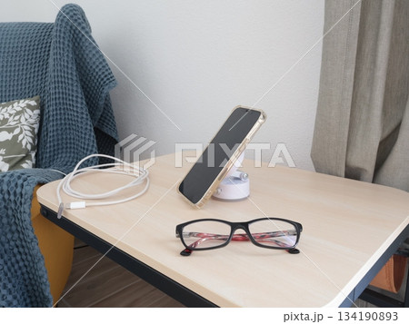 Holder, tabletop stand for smartphone and glasses on table near armchair. Modern device at home. Technology and age. Holder, tabletop stand for smartphone and glasses on table near armchair. Modern device at home. Technology and age. 134190893