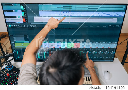 Top view of sound engineer male creating new music in home studio, arranging tracks and mixing audio levels with midi keyboard and software on a large display, showcasing modern music production. 134191193