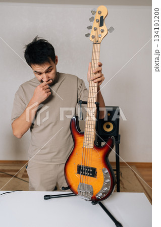 Vertical portrait of male musician, standing alongside bass guitar on stand, deeply concentrating and thinking about music composition, exploring inspiration, pursuing creative ideas in studio. 134191200