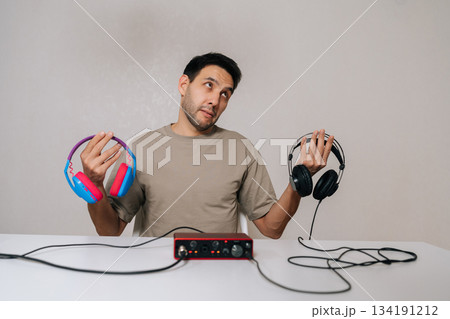 Cheerful man weighing colorful expressive headphones against black professional studio pair tied to audio interface, pondering audio quality, style and creative vs technical choice. 134191212