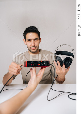 Vertical portrait of man holding audio interface and headphones, demonstrating setup for sound recording or music production with hand offering cable connection sitting at white table looking camera. 134191220