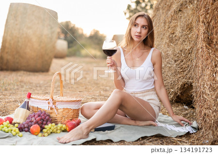 Young pretty woman in white blouse and shorts sitting on soft mat near stack of straw with glass of wine, romantic picnic in field Young pretty woman in white blouse and shorts sitting on soft mat near stack of straw with glass of wine, romantic picnic in field 134191723