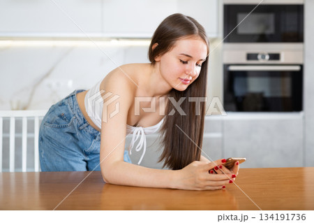 Interested girl standing in home kitchen, browsing through websites or chatting on social media on smartphone while leaning on table Interested girl standing in home kitchen, browsing through websites or chatting on social media on smartphone while leaning on table 134191736