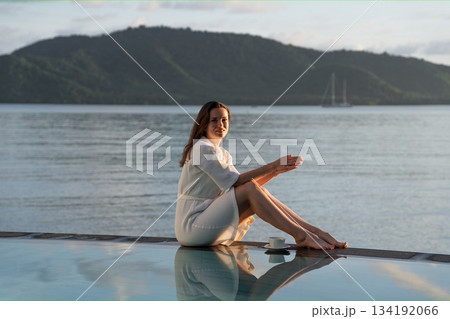 brunette white bathrobe sits at edge of pool overlooking Indian Ocean 134192066