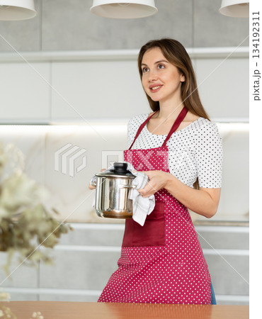 Portrait of positive housewife in an apron with pot of soup in her hands Portrait of positive housewife in an apron with pot of soup in her hands 134192311