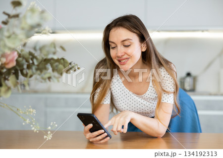 Middle-aged woman looking at her phone leaning on kitchen table Middle-aged woman looking at her phone leaning on kitchen table 134192313