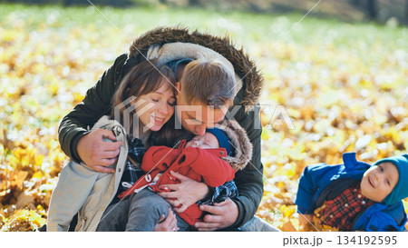 Parents cuddling their sleeping baby in a park full of fall foliage while their older son plays in the leaves Parents cuddling their sleeping baby in a park full of fall foliage while their older son plays in the leaves 134192595