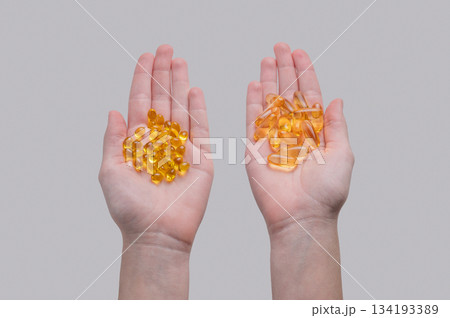 Hands holding two types of yellow softgel capsules containing D3 and Omega on a light background. Hands holding two types of yellow softgel capsules containing D3 and Omega on a light background. 134193389