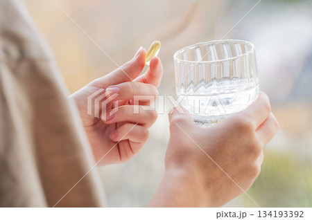 A close-up of a person's hands holding yellow softgel in one palm and a glass of water in the other. A close-up of a person's hands holding yellow softgel in one palm and a glass of water in the other. 134193392