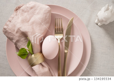 Easter table setting with pastel pink egg, gold cutlery and linen napkin on soft pink plate. Elegant spring decor. 134195534