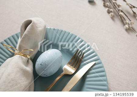 Elegant Easter table setting with blue speckled egg, gold cutlery and linen napkin on pastel plate and linen tablecloth. Soft spring decor and holiday atmosphere. 134195539