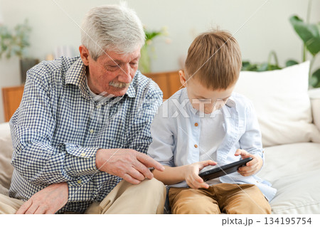 Happy family at home. Old senior man grandfather boy grandson enjoying time together watching video on phone. Good time at home. grandparent and child together. Older generation modern tech usage Happy family at home. Old senior man grandfather boy grandson enjoying time together watching video on phone. Good time at home. grandparent and child together. Older generation modern tech usage 134195754