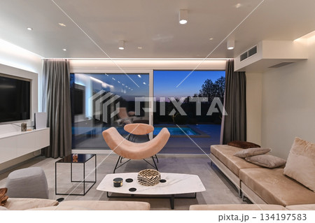 Elegant modern living room with designer lounge chair, panoramic glass doors, sunset view, and poolside terrace in a luxury contemporary home interior Elegant modern living room with designer lounge chair, panoramic glass doors, sunset view, and poolside terrace in a luxury contemporary home interior 134197583
