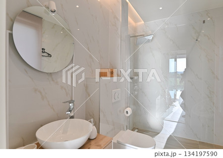 Modern, elegant bathroom featuring white marble-look tiles, a wooden vanity with a vessel sink, a wall-mounted toilet, and a glass shower, illuminated by sleek ambient lighting 134197590