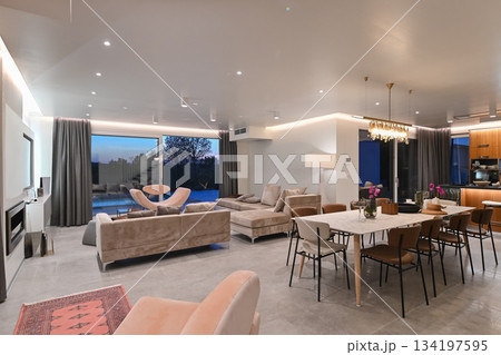 Modern luxury living room interior at sunset. Open-plan space featuring beige sofas, marble dining table, large glass windows with sun flare, and view of a patio. Stylish contemporary home design Modern luxury living room interior at sunset. Open-plan space featuring beige sofas, marble dining table, large glass windows with sun flare, and view of a patio. Stylish contemporary home design 134197595