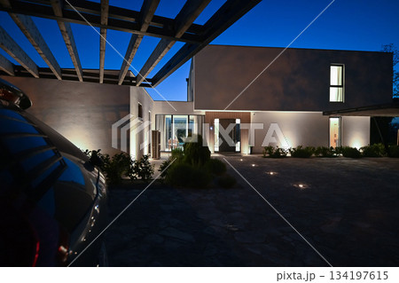 Exterior of a modern luxury house at night viewed from a carport. Illuminated white villa with paved driveway, wooden pergola, and car silhouette against a deep blue twilight sky 134197615
