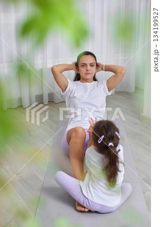 Mother doing crunches on a yoga mat with her daughter watching nearby 134199527