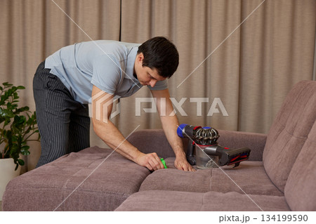 Man cleaning sofa upholstery at home using a powerful handheld vacuum cleaner 134199590