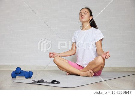 Woman sitting in lotus pose on a mat, meditating after exercise session at home Woman sitting in lotus pose on a mat, meditating after exercise session at home 134199647