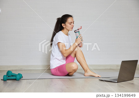 Young woman hydrating with a water bottle after exercising, using a laptop for online fitness 134199649