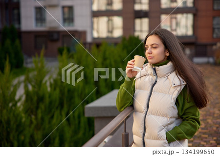 Woman standing on a modern park, drinking coffee and looking thoughtful 134199650