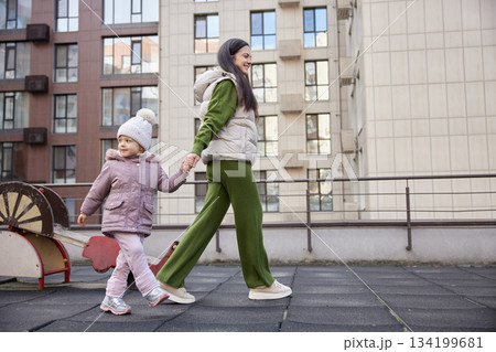 Mother and daughter holding hands, walking together across an urban park playground Mother and daughter holding hands, walking together across an urban park playground 134199681