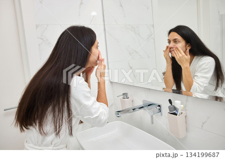 woman applying skincare cream to her face in the bathroom mirror 134199687