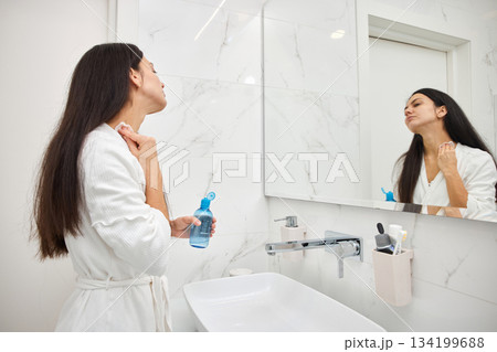 Woman cleansing her face with toner and cotton pad looking in bathroom mirror 134199688