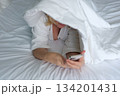 Smiling woman lying on a bed with a blanket over her head while using the mobile phone 134201431