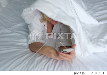 Smiling woman lying on a bed with a blanket over her head while using the mobile phone Smiling woman lying on a bed with a blanket over her head while using the mobile phone 134201431