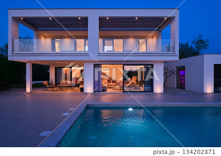 Exterior view of a modern luxury house with a swimming pool at twilight. Illuminated two-story white villa with large glass windows, patio, terrace, and glowing blue water Exterior view of a modern luxury house with a swimming pool at twilight. Illuminated two-story white villa with large glass windows, patio, terrace, and glowing blue water 134202871