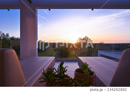 Sunset view from a luxurious modern patio with lounge chairs, overlooking a tranquil green landscape of trees and vineyards, perfect for a relaxing evening Sunset view from a luxurious modern patio with lounge chairs, overlooking a tranquil green landscape of trees and vineyards, perfect for a relaxing evening 134202892