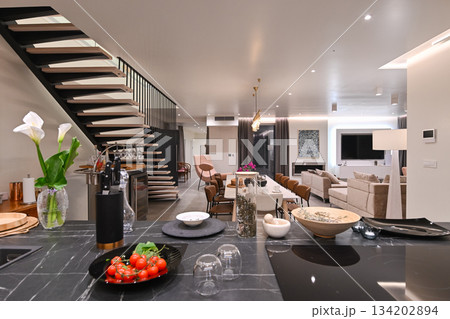Modern luxury open-plan interior featuring a floating staircase, marble dining table, wooden kitchen, and beige sofa. Spacious contemporary home design with elegant lighting and gray tiled floors Modern luxury open-plan interior featuring a floating staircase, marble dining table, wooden kitchen, and beige sofa. Spacious contemporary home design with elegant lighting and gray tiled floors 134202894