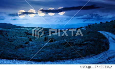 country dirt road through mountains on a summer night. scenic view of rolling hills under dark sky with clouds in full moon light. beautiful alpine meadow for finance investment or abstract concepts 134204502