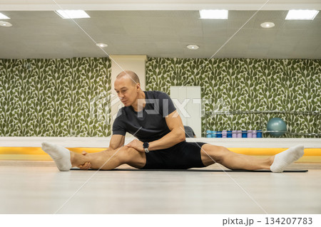 Adult caucasian male doing wide-leg seated stretch on mat, improving flexibility and posture in fitness studio. 134207783