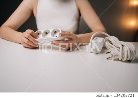 Cropped shot of unrecognizable woman gripping white triple electrical socket sitting at white desk, highlighting electrical work, home improvement, energy efficiency in dark interior setting Cropped shot of unrecognizable woman gripping white triple electrical socket sitting at white desk, highlighting electrical work, home improvement, energy efficiency in dark interior setting 134208721