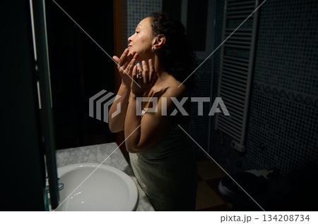 Side view of a beautiful young adult woman 40s, applying hydrating moisturizing face cleaning cosmetic product, standing in front of a bathroom mirror, wrapped in bath towel. 134208734