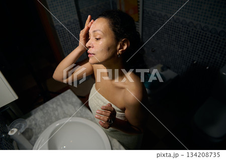 Confident portrait of a middle aged pretty woman applying cosmetic product, standing in front of a bathroom mirror, wrapped in bath towel, enjoying daily morning routine and procedures 134208735