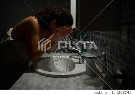 Authentic portrait of a woman washing her face. Drops of water splashing over a ceramic sink. Shot with low key light 134208745