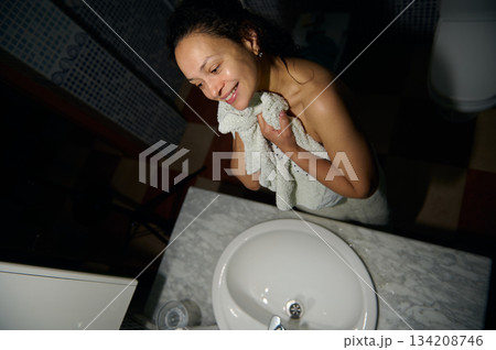 Smiling woman wrapped with a bath towel, wiping her face, standing by a white ceramic sink in the bathroom Smiling woman wrapped with a bath towel, wiping her face, standing by a white ceramic sink in the bathroom 134208746