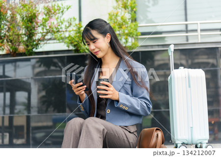 Woman business person lifestyle travel street work outdoor, stylish young professional enjoying coffee, using smartphone, on journey break, light blue suitcase and brown bag nearby. Woman business person lifestyle travel street work outdoor, stylish young professional enjoying coffee, using smartphone, on journey break, light blue suitcase and brown bag nearby. 134212007