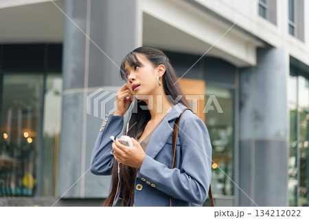 Travel woman, young person enjoying business lifestyle, inserts earphones for work on bustling city street, vibrant outdoor urban setting perfect for modern professionals. 134212022