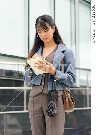 Young woman on city street, business professional lifestyle person working outdoor writing notes in book carrying umbrella shoulder bag standing urban city background focused on task. Young woman on city street, business professional lifestyle person working outdoor writing notes in book carrying umbrella shoulder bag standing urban city background focused on task. 134212036