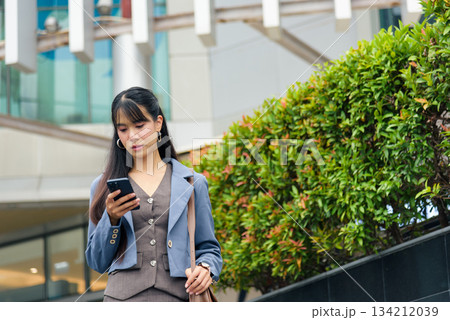 Young corporate woman walks urban street uses smartphone for work communication outdoor in busy city environment with modern building and green foliage behind her. Young corporate woman walks urban street uses smartphone for work communication outdoor in busy city environment with modern building and green foliage behind her. 134212039