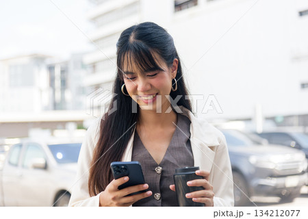 Smiling young business woman, engaged person enjoying modern urban lifestyle, using smartphone, holding coffee cup, walking outdoor city street, representing work and travel. 134212077