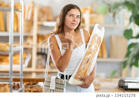 Young woman holding baguettes looks around the bakery Young woman holding baguettes looks around the bakery 134215278
