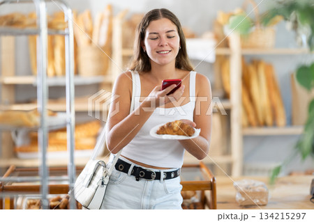 Young woman stands in bakery, scanning a croissant with her phone 134215297
